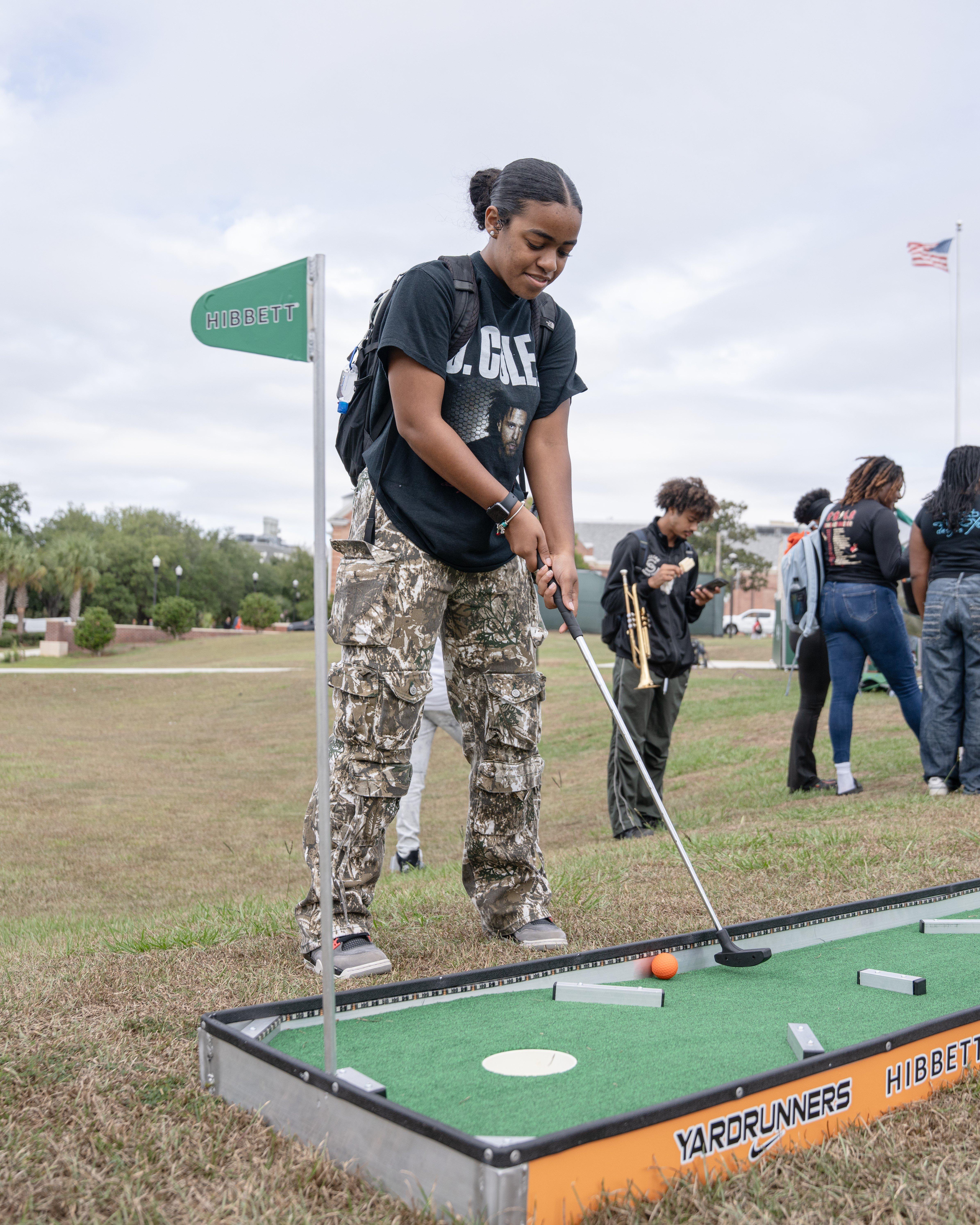 A stylish woman stands next to a vintage green muscle car, which is decorated with flowers and features the FAMU snake mascot design on the door. She is wearing a cream-colored t-shirt with the snake design and a black skirt.